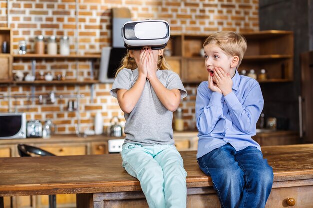 Hermanos pequeños sorprendidos y alegres probando el dispositivo VR mientras están sentados en la cocina