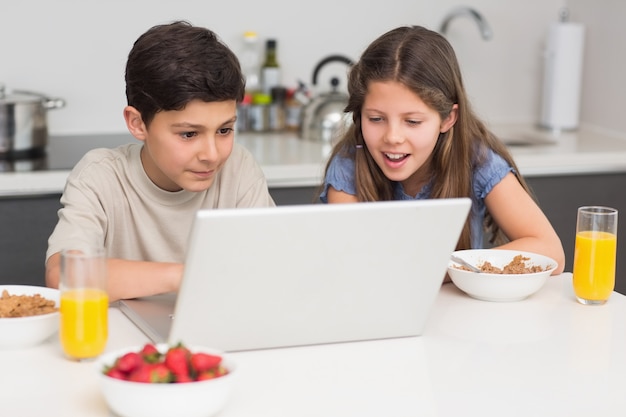 Hermanos sonrientes que disfrutan del desayuno mientras que computadora portátil en cocina