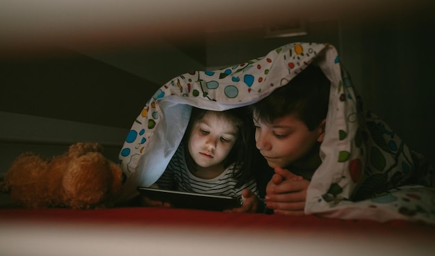 Hermanos usando una tableta digital bajo la manta en casa