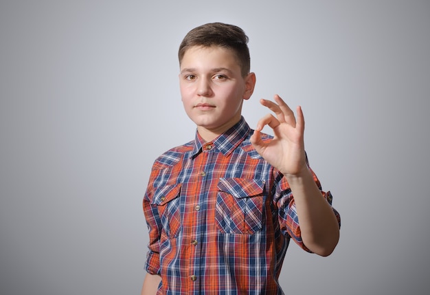 Hermosa adolescente con una camisa a cuadros, mostrando el signo de OK en la superficie gris-blanca