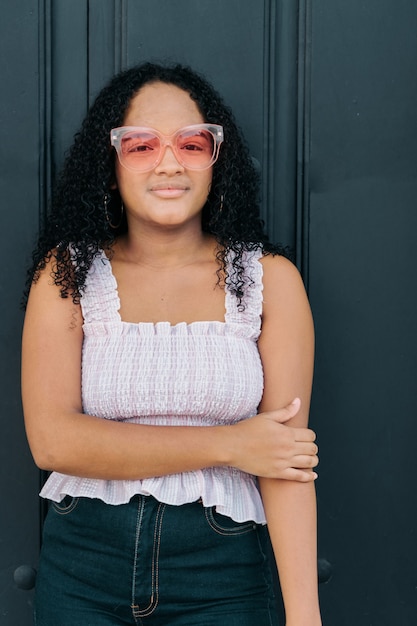 Hermosa adolescente colombiana con gafas de sol de pie frente a la puerta de madera.