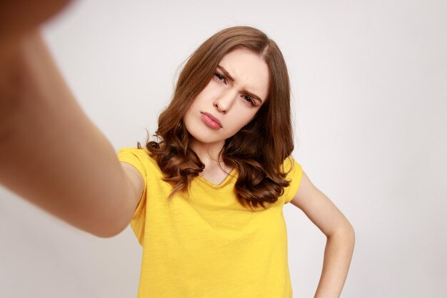 Hermosa adolescente confundida con una camiseta informal amarilla mirando la cámara POV, punto de vista de la foto, cara fruncida, expresión de malentendidos. Disparo de estudio interior aislado sobre fondo gris.