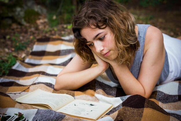 Hermosa adolescente leyendo un libro sobre una manta sobre hierba verde en el bosque.