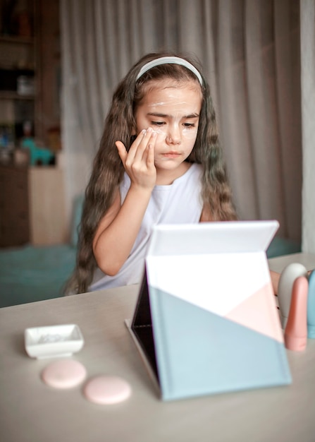 Hermosa adolescente viendo la clase magistral de belleza con tableta en línea y haciendo el procedimiento de spa por sí misma, niño con máscara suave en la cara, salón de belleza en casa