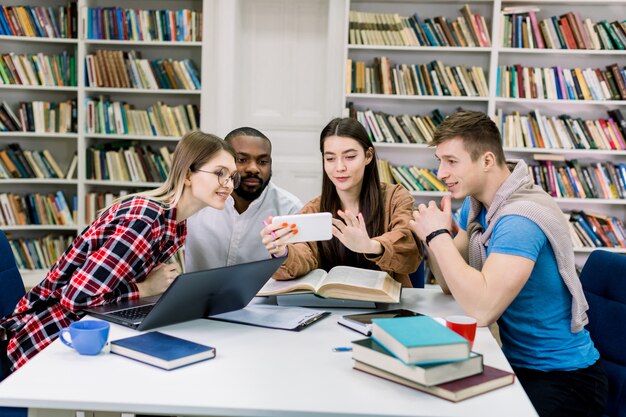 Hermosa alegre joven estudiante caucásica mostrando información en su teléfono inteligente a sus alegres amigos multiétnicos durante la preparación de su equipo para los exámenes en la biblioteca