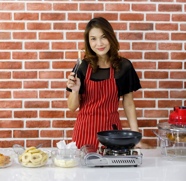 Hermosa ama de casa asiática en delantal de rayas rojas sonriendo felizmente mientras disfruta cocinando papas fritas con sartén negra en la mesa de la cocina cerca de la pared de ladrillo que se llena con utensilios de cocina, cuencos de vidrio e ingredientes