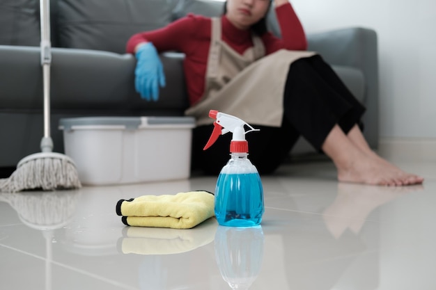 La hermosa ama de casa asiática se siente cansada después de hacer las tareas domésticas se sienta en el suelo de la sala de estar con productos de limpieza para el hogar