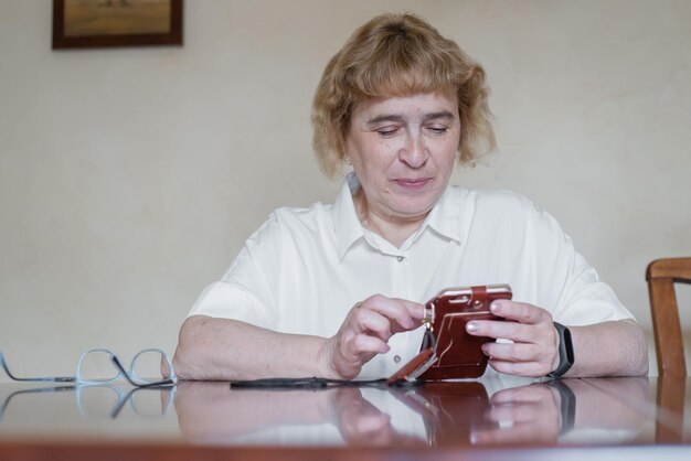 Hermosa anciana con una blusa blanca sentada en la mesa y escribe mensajes en el teléfono Mujer jubilada anotó puntos y lee el mensaje en el teléfono inteligente