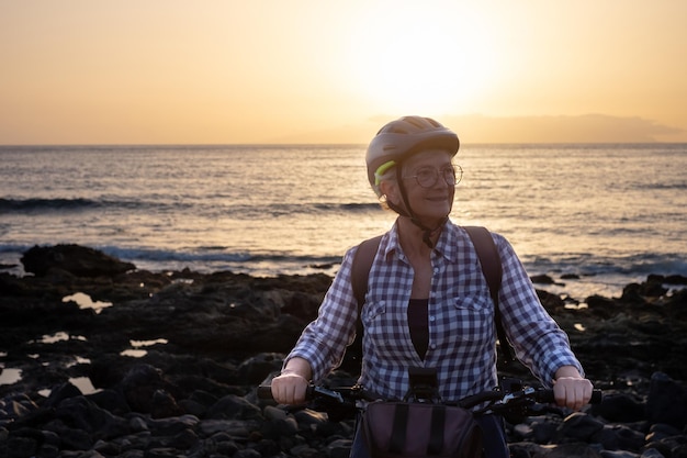 Hermosa anciana caucásica activa con casco de ciclismo mientras monta en bicicleta al atardecer en la playa anciana disfrutando de un estilo de vida saludable