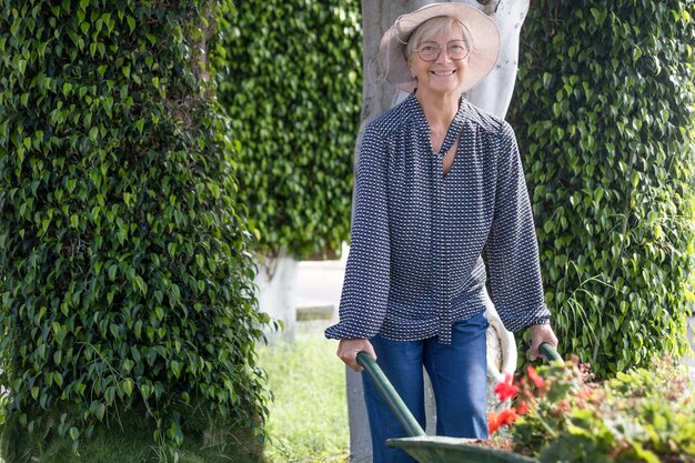 Hermosa anciana caucásica jardinería sosteniendo una carretilla llena de flores Fondo verde Amor por el concepto de naturaleza