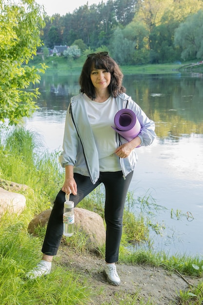 Hermosa anciana se encuentra en la orilla del río con una alfombra de yoga y una botella de agua