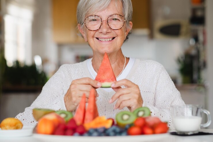 Hermosa anciana feliz sosteniendo una rodaja de sandía mientras desayuna en casa con leche de fruta fresca de temporada y concepto de alimentación saludable de cupcakes