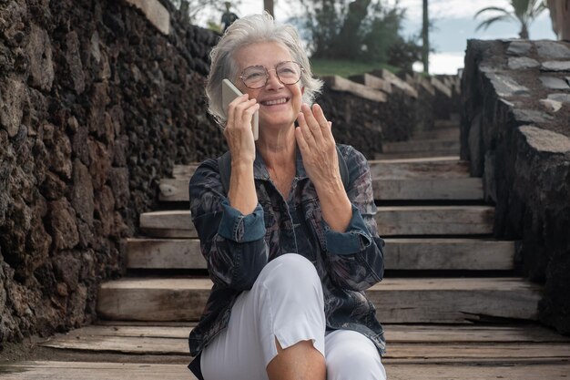 Hermosa anciana sentada al aire libre en una escalera de madera mientras habla por teléfono móvil Feliz anciana sonriente usando tecnología inalámbrica