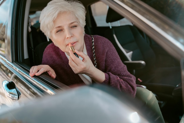 Hermosa anciana sentada en el asiento delantero del vehículo de motor aplicando el lápiz labial en su labio superior