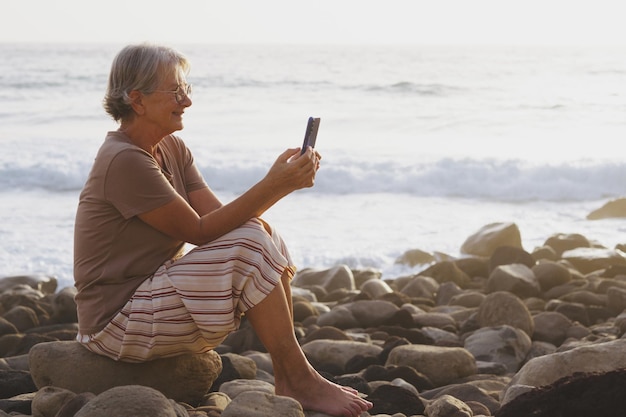 Hermosa anciana sentada en la playa al atardecer mirando su teléfono inteligente sonriendo anciana relajada disfruta de vacaciones y libertad