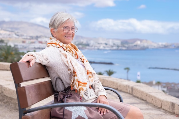 Hermosa anciana sonriente con anteojos sentada al aire libre en un banco mirando el mar disfrutando de la libertad y las vacaciones Anciana activa relajándose en un día soleado