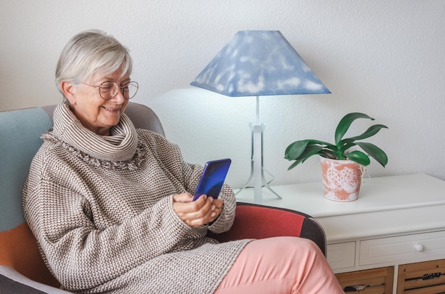 Hermosa anciana sonriente sentada en casa usando un teléfono móvil disfrutando de la tecnología y las redes sociales de la vieja generación y el nuevo concepto de tecnología