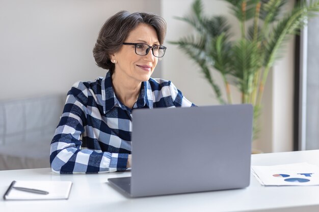 Hermosa anciana trabajando de forma remota en casa en internet usando laptop