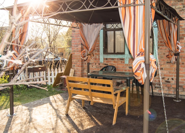Una hermosa área de recreación para una familia en una casa de campo, una carpa con un banco y una mesa en el fondo de una casa de ladrillos con ventanas y techo, primavera, puesta de sol