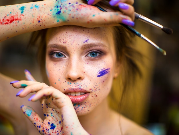 hermosa artista femenina con manos sucias con diferentes pinturas sobre ellas, sosteniendo pinceles cerca de su cara