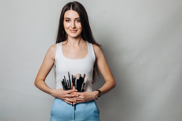 Hermosa artista femenina con pinceles de maquillaje en salón de belleza en una pared de luz