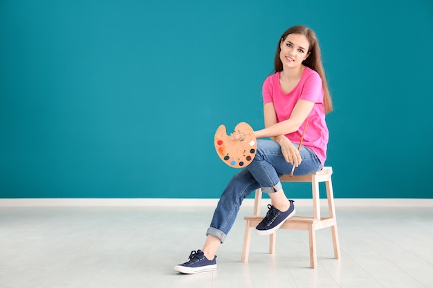 Hermosa artista femenina sentada en un taburete contra la pared de color