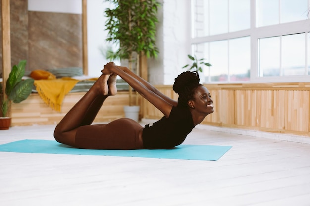 Hermosa y atlética mujer afroamericana acostada en la pose de proa vista lateral haciendo yoga en una colchoneta de gimnasia en un estudio decorado Músculo estirando ejercicios gimnásticos fitness manteniendo el cuerpo en forma