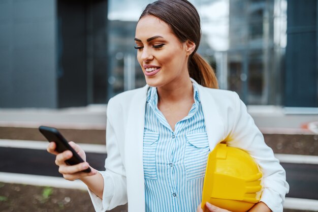 Hermosa atractiva arquitecta caucásica de pie frente a su empresa con casco debajo de la axila y usando un teléfono inteligente para leer el correo electrónico.