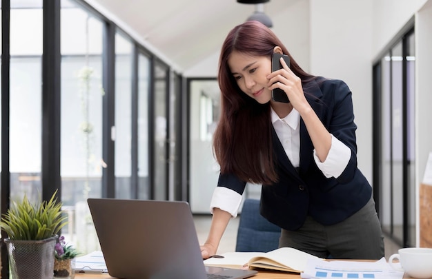 Hermosa y atractiva joven empresaria asiática hablando por teléfono con su colega y revisando la información en la pantalla del portátil