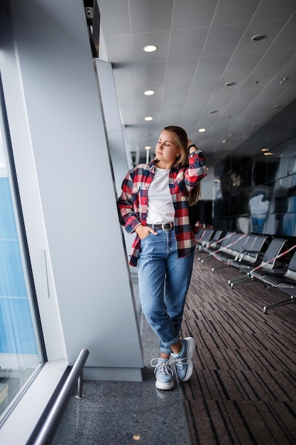 Una hermosa y atractiva mujer ucraniana con cabello rubio está de pie junto a una gran ventana usando jeans momom y una camiseta con una camisa a cuadros Chica con ropa casual y cómoda