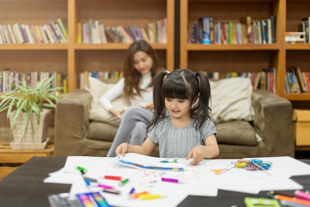 Hermosa y atractiva niña caucásica haciendo pintura con la madre lee el libro detrás del concepto de ideas familiares de la casa