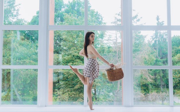 Hermosa bailarina con ropa hermosa se para en el alféizar de la ventana y sostiene una canasta de flores