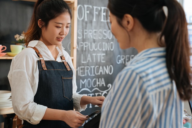 Hermosa barista femenina en delantal tomando tarjeta de crédito y usando tableta mientras trabaja en el mostrador de la cafetería. cliente de la señora de la oficina esperando el cheque en la cafetería moderna. cliente pagando con tarjeta de debito