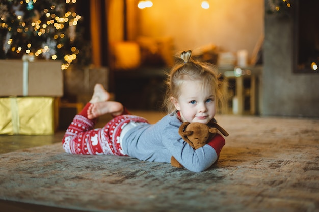 Una hermosa bebe yace en el suelo frente al árbol y abraza a su peluche favorito