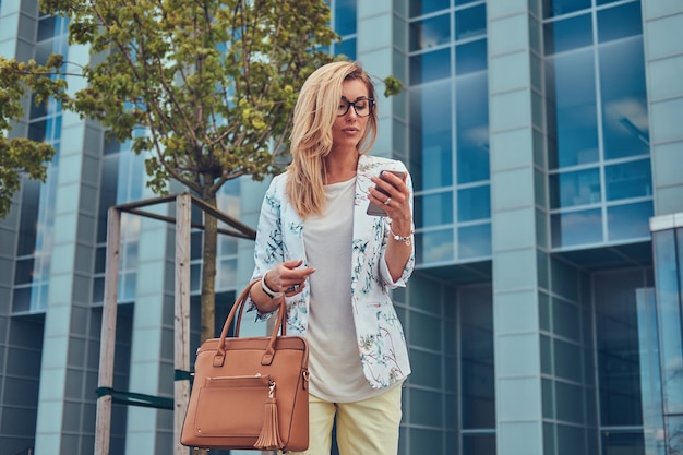 Hermosa bloguera de moda con ropa elegante y gafas con un bolso, sostiene un teléfono inteligente, de pie contra un rascacielos.
