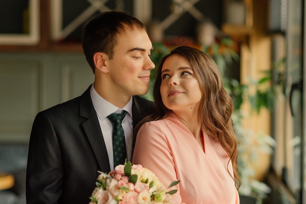 Hermosa boda, esposo y esposa, novia y novio de pie en el desván interior junto a la ventana. Pareja de recién casados enamorados. Novio abraza a la novia por los hombros.