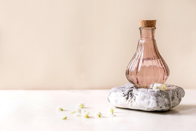 Hermosa botella rosa transparente de perfume con corcho sobre piedra texturizada sobre una mesa de mármol blanco con pequeñas flores blancas Kalanchoe alrededor Concepto de fabricación de perfume con lugar para la etiqueta de diseño