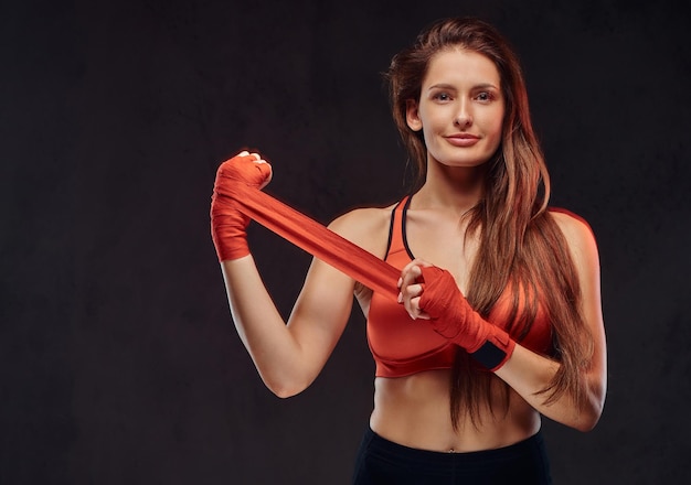 Una hermosa boxeadora morena sonriente con sostén deportivo venda sus manos. Aislado sobre fondo oscuro con textura.