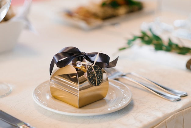 Una hermosa caja de papel de oro con un lazo de satén marrón, un bombón de boda, en un plato blanco sobre la mesa del banquete.