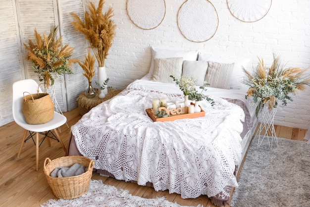 Hermosa casa interior en tonos blancos y beige, con atrapasueños, flores secas y una cama. Interior de una casa acogedora