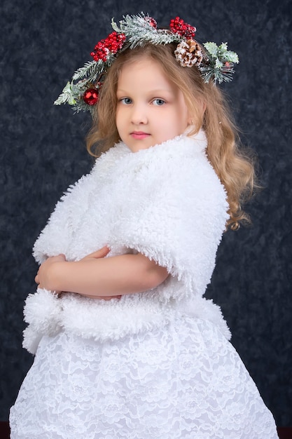 Hermosa chica en un abrigo de piel blanco con una corona de Navidad en la cabeza sobre un fondo gris