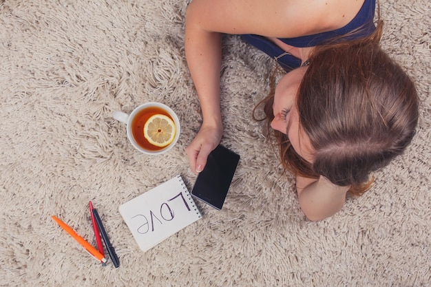 Hermosa chica acostada en la alfombra en casa y leyendo sms