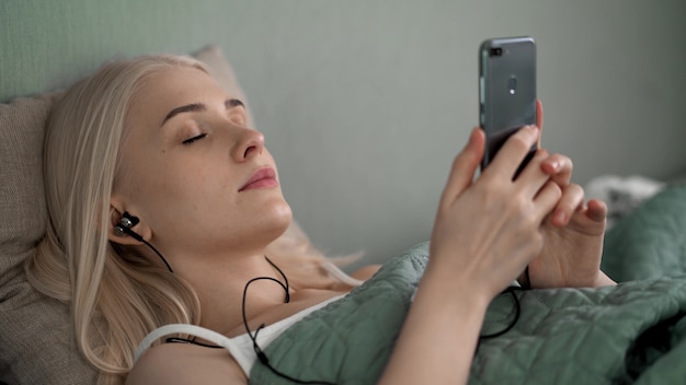 Hermosa chica acostada en la cama escuchando música con auriculares. Pasa tu tiempo libre en casa.