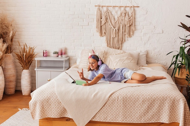 Hermosa chica adolescente se comunica en línea por teléfono mientras está acostada en la cama en casa. Niño en auriculares aprende en línea mientras está acostado en la cama. Concepto de aprendizaje a distancia