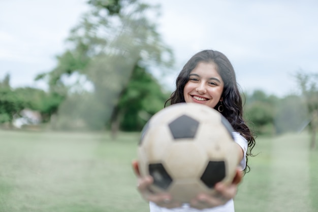 Hermosa chica adolescente sosteniendo un balón de fútbol y borrosa fondo de naturaleza verde