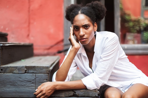 Hermosa chica africana con cabello rizado oscuro en camisa blanca mirando cuidadosamente a la cámara mientras se apoya en las escaleras en el viejo patio