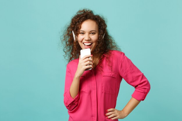 Hermosa chica africana en ropa casual sostiene el teléfono móvil, escucha música con auriculares aislados sobre fondo azul turquesa en estudio. Concepto de estilo de vida de emociones sinceras de personas. Simulacros de espacio de copia.