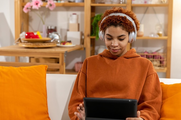 Hermosa chica afroamericana con peinado afro usando auriculares sonriendo escuchando música en casa interior Mujer joven disfruta escuchando música favorita en tableta Concepto de alegría de estilo de vida de personas