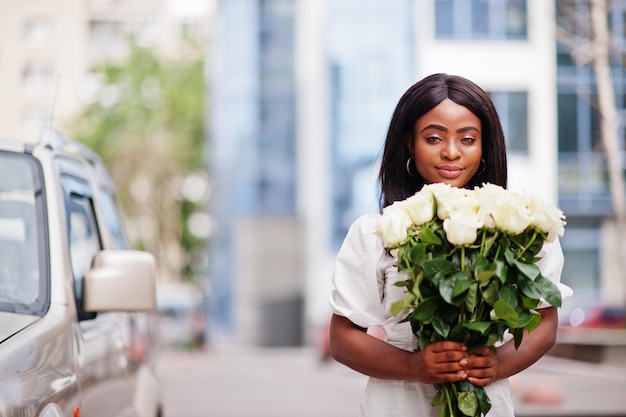 Hermosa chica afroamericana con ramo de flores de rosas blancas en citas en la ciudad. Mujer de negocios negra con ramo de flores cerca del auto.