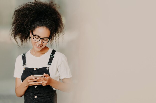 Hermosa chica afroamericana con ropa de moda usa el teléfono móvil para enviar mensajes de texto conectados a wifi comprueba el buzón de correo electrónico se encuentra cerca de la pared blanca con espacio vacío Comunicación en línea